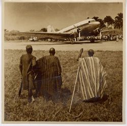 Three men in traditional dress watch airplane and crowd from distance