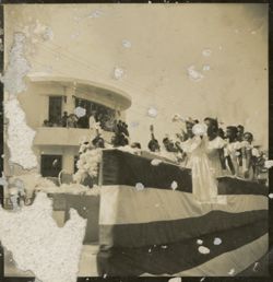 Women in white dresses wave to officials on balcony while riding on float in military parade outside of Executive Pavilion