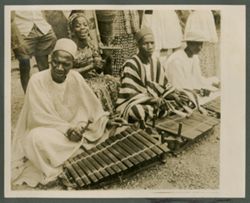 Men in traditional attire playing instruments while sitting on ground