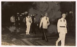 Men in military uniforms and decorated formal wear walk together in procession