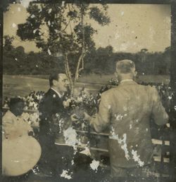 One dignitary hands another a case while standing on a balcony in front of a crowd