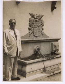 Dignitary stands beside a fountain decorated with a Spanish coat of arms