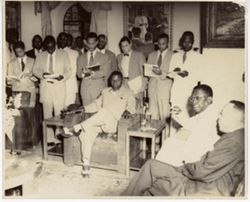 President Tubman converses with seated companions while group of men take notes