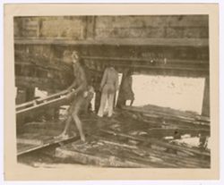 Man carries a pallet of wood in the foreground while a man and a young women stand under a pier or bridge