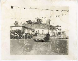 Men ride on outside of car during a parade with crowds gathered