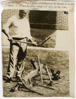 Writer holding a grass cutter poses by a plow with an African ax on the handle