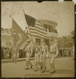 Men walk in parade holding American and Marines flags in front of Executive Pavilion
