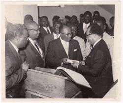 Vice President Tolbert, President Tubman, and Nicolas Grunitzky stand with other officials behind a large book on a podium