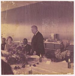 Man stands at banquet table while President William V. S. Tubman and others look on
