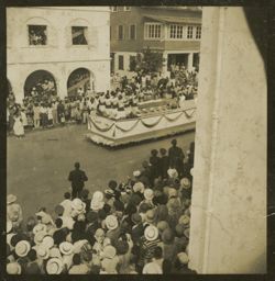 Women in matching dresses ride a parade float past crowded sidewalks