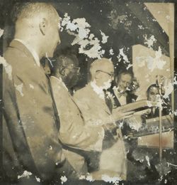 Man surrounded by other dignitaries, including President Tubman, holds a book