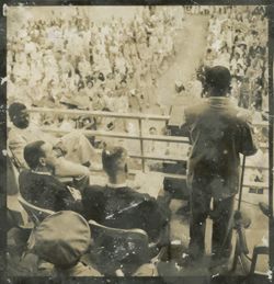 President Tubman stands on a balcony facing a crowd, digitaries and military officers are seated around him