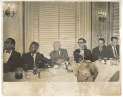 Men in business suits sit at table and look toward head of table