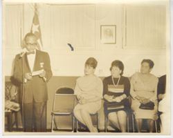 Consul General Thomas speaks into microphone while Mrs. Thomas, Mrs. Leonard Epstein and Mrs. Isabelle Edwin listen