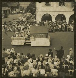 Women in coordinating dresses ride a parade float with a cabin on it