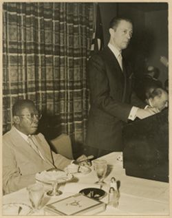 President William V. S. Tubman seated at table next to man standing at podium