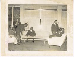 Men sit and stand together at chairs in semicircle around table