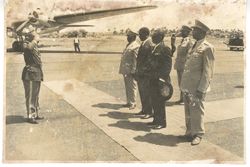 Man in military dress salutes to President William V. S. Tubman and others at airport