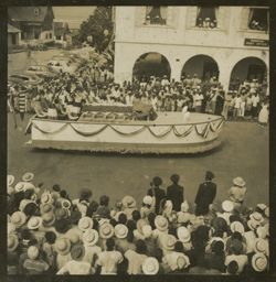 Women in matching dresses stand on a boat shaped parade float past a crowd