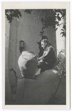 Item 0527. Alexandrov seated on an adobe balustrade beside a large jug with legs and handles.