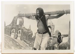 Item 0193.Two Indigenous men wearing black cloth over their heads and no shirts. Their outstretched arms are tied with rope to a long cactus stem. Taken on the roof of a church, the towers of which are seen in the background.