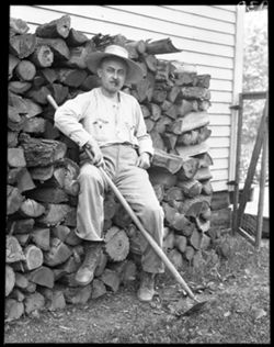 Richard Lieber with hoe on woodpile, seated