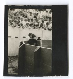 Item 0980. Alexandrov with camera behind inner fence of arena. Head of matador visible in center foreground. Spectators in upper background.