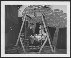 Hoagy Carmichael on soundstage sitting in a swing with an unidentified girl during the taping of the Saturday Night Revue television program, 1954 or 1955.
