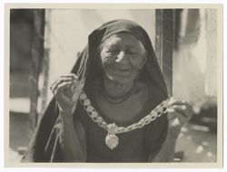 Close-up of an elderly Indigenous woman wearing a dark cloth veil on her head and holding up a chain and coin (?) necklace. Her eyes are closed and she is smiling.