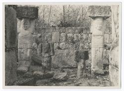 Item 0181. Two large and two small stone statues enclosed in a small walled area, possibly at the Southeast Colonnade. The two larger figures support large stone blocks on their heads.