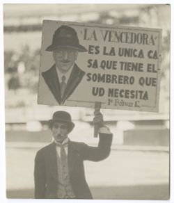 Item 0146. Man in Chaplinesque moustache and outfit, holding up large sign on a stick. Sign has a picture of a man in a hat, and the words: "La Vencedora'/Es la unica ca-/sa que tience el/sombrero que/ud. Necesita/la Bolivar"