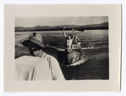 Item 1175. On a lake (?) with mountains in far background. Man wearing a soft hat stands behind a large, square object draped with a cloth. Behind him, in a canoe, a man is holding a paddle over his head, and behind him, a second canoe is visible in which a man is seated with his back to the camera.