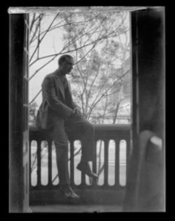 Item 0273. Unidentified man in business suit sitting on balcony railing. Trees, street or park in background.