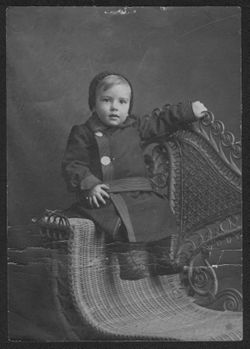 Portrait of Hoagy Carmichael as a little boy in his winter coat sitting on the arm of a wicker chair.