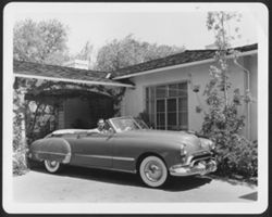 Hoagy Carmichael and Ruth Carmichael sitting in a large Oldsmobile convertible in the driveway of a house.