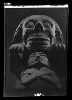 Item 0260. Probably taken at National Museum of Anthropology. Large unidentified stone statue with large earring and prominent teeth. Small stone figure with crossed arms in front.
